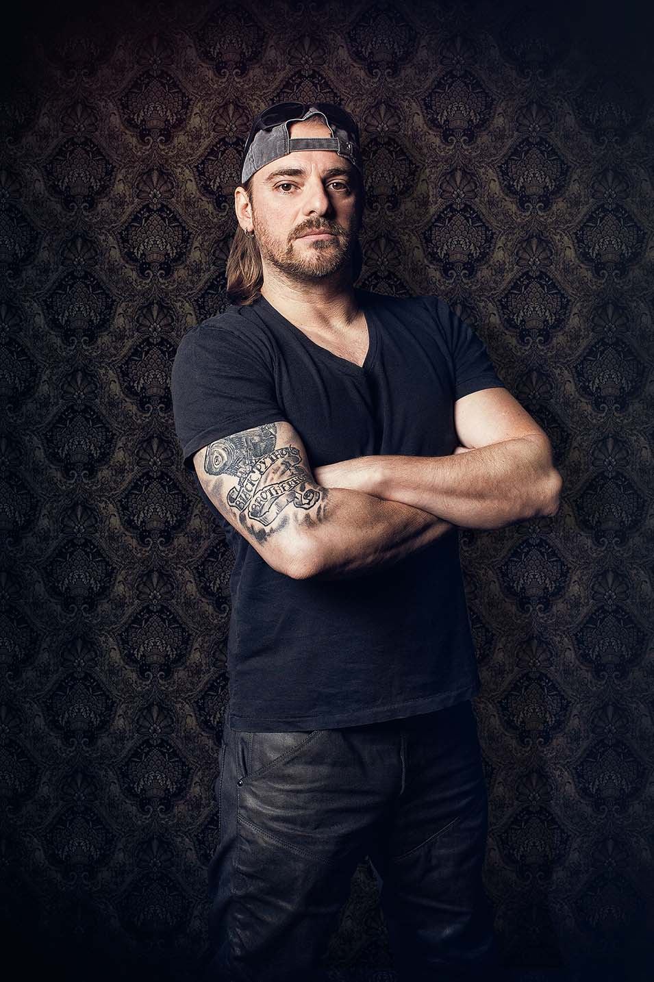 Oliver Sasin, Inhaber und Tätowierer von LE Ink, mit verschränkten Armen vor dunkler Tapete